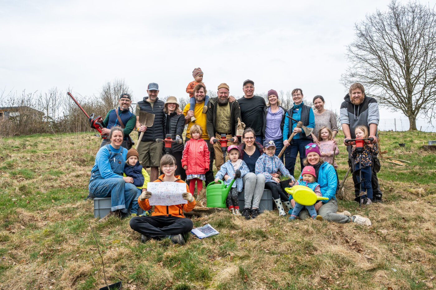 Eine große Gruppe von Familien mit Kindern und Werkzeugen bei einer gemeinschaftlichen Baumpflanzaktion auf einer Wiese.