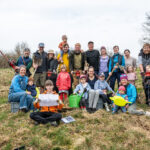 Eine große Gruppe von Familien mit Kindern und Werkzeugen bei einer gemeinschaftlichen Baumpflanzaktion auf einer Wiese.